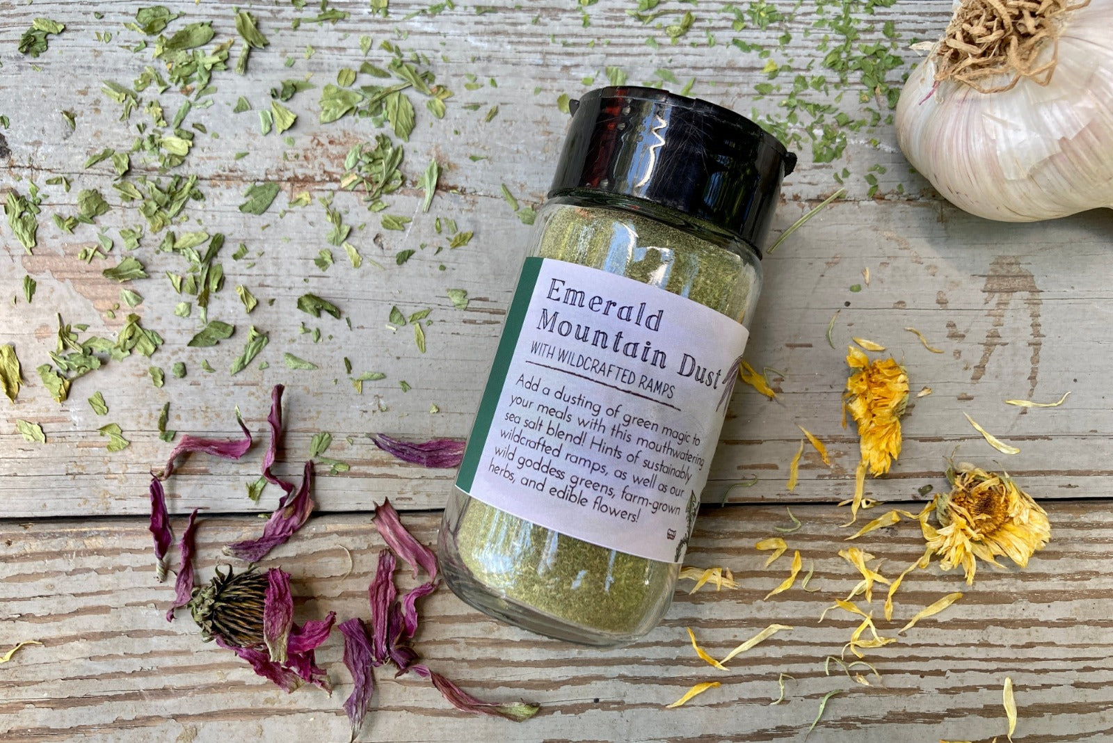 A jar of Emerald Mountain Dust from Well Seasoned Table on a wooden background with a sprinkle of calendula flowers and dried ramps.
