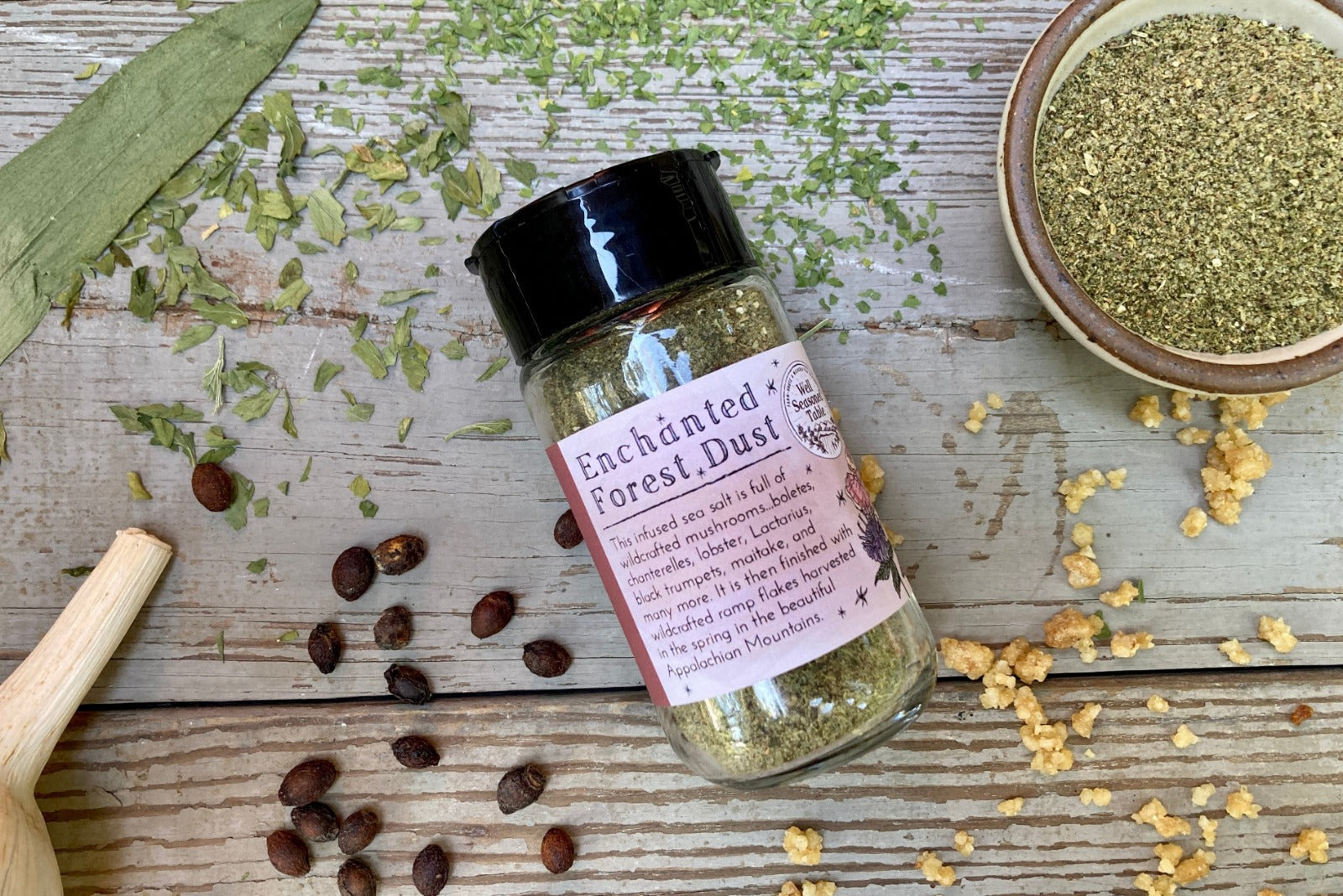 A glass shaker jar of Enchanted Forest dust from Well Seasoned Table, a wild mushroom and herb infused sea salt, on a wooden background with garlic, herbs, ramps, and spicebush berries.