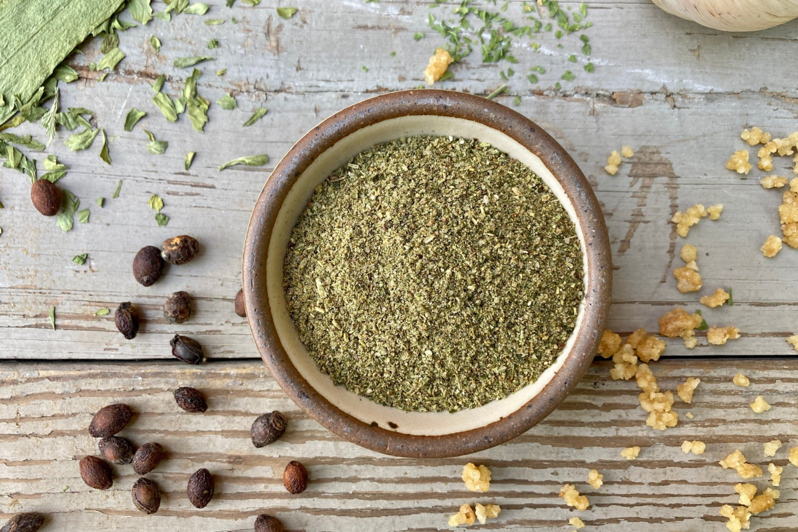A glass shaker jar of Enchanted Forest dust from Well Seasoned Table, a wild mushroom and herb infused sea salt, on a wooden background with garlic, herbs, ramps, and spicebush berries.