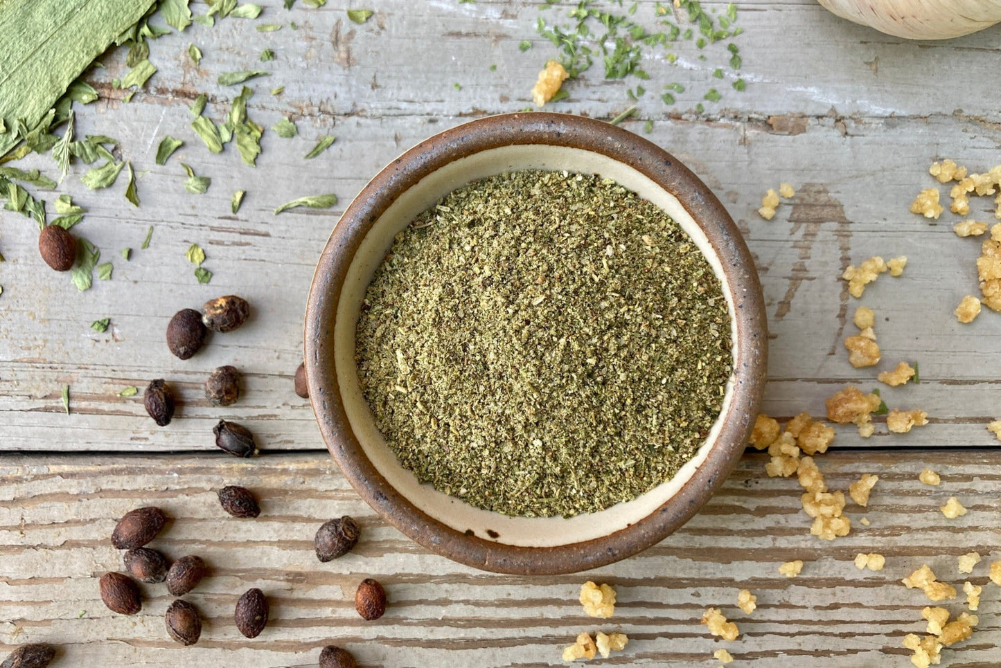 A glass shaker jar of Enchanted Forest dust from Well Seasoned Table, a wild mushroom and herb infused sea salt, on a wooden background with garlic, herbs, ramps, and spicebush berries.