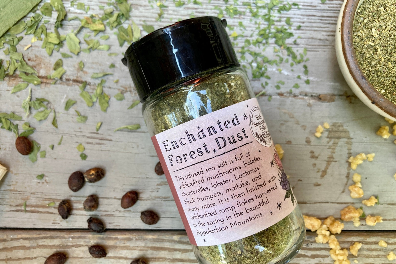 A glass shaker jar of Enchanted Forest dust from Well Seasoned Table, a wild mushroom and herb infused sea salt, on a wooden background with garlic, herbs, ramps, and spicebush berries.