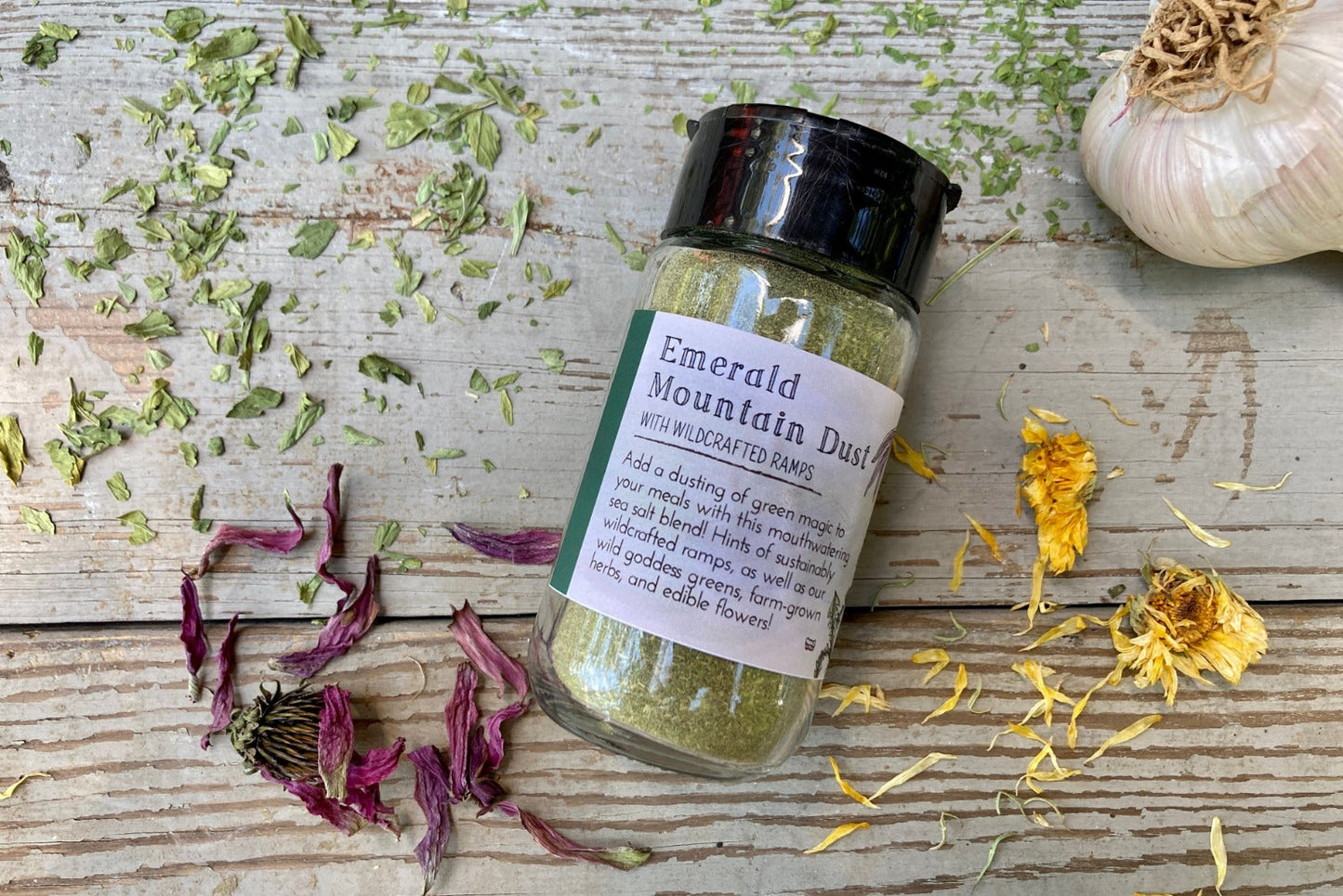 A jar of Emerald Mountain Dust from Well Seasoned Table on a wooden background with a sprinkle of calendula flowers and dried ramps. 