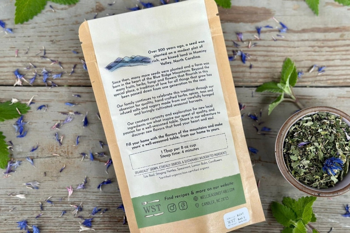 The back of a packet of in the garden with tulsi tea from Well Seasoned Table on a wooden background with fresh spearmint and cornflowers around it and a ceramic bowl of tea.