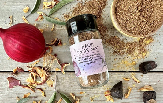 A glass seasoning jar of Magic Onion Dust from Well Seasoned Table on a wooden background with onions, sage, and black garlic around it.