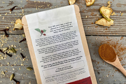 The back of a packet of Bourbon Spiced Apple Pie Chai Tea from Well Seasoned Table on a wooden background with dried apples, spices, and a spoon of cinnamon around it.