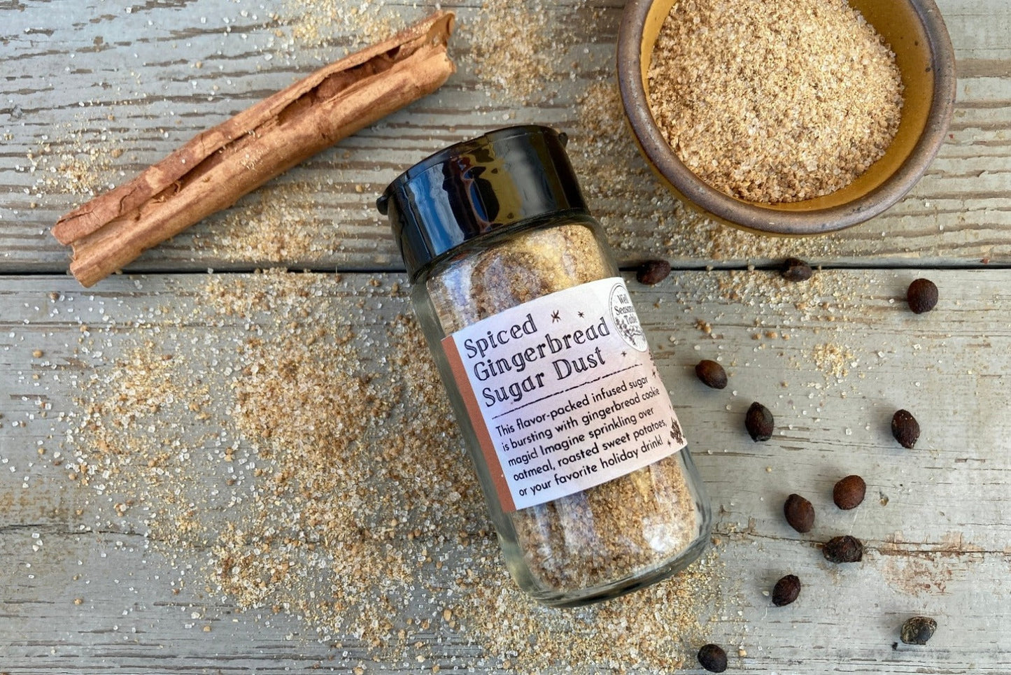 A glass shaker jar of Spiced Gingerbread Sugar Dust from Well Seasoned Table on a wooden background with a bowl of gingerbread sugar and a cinnamon stick.