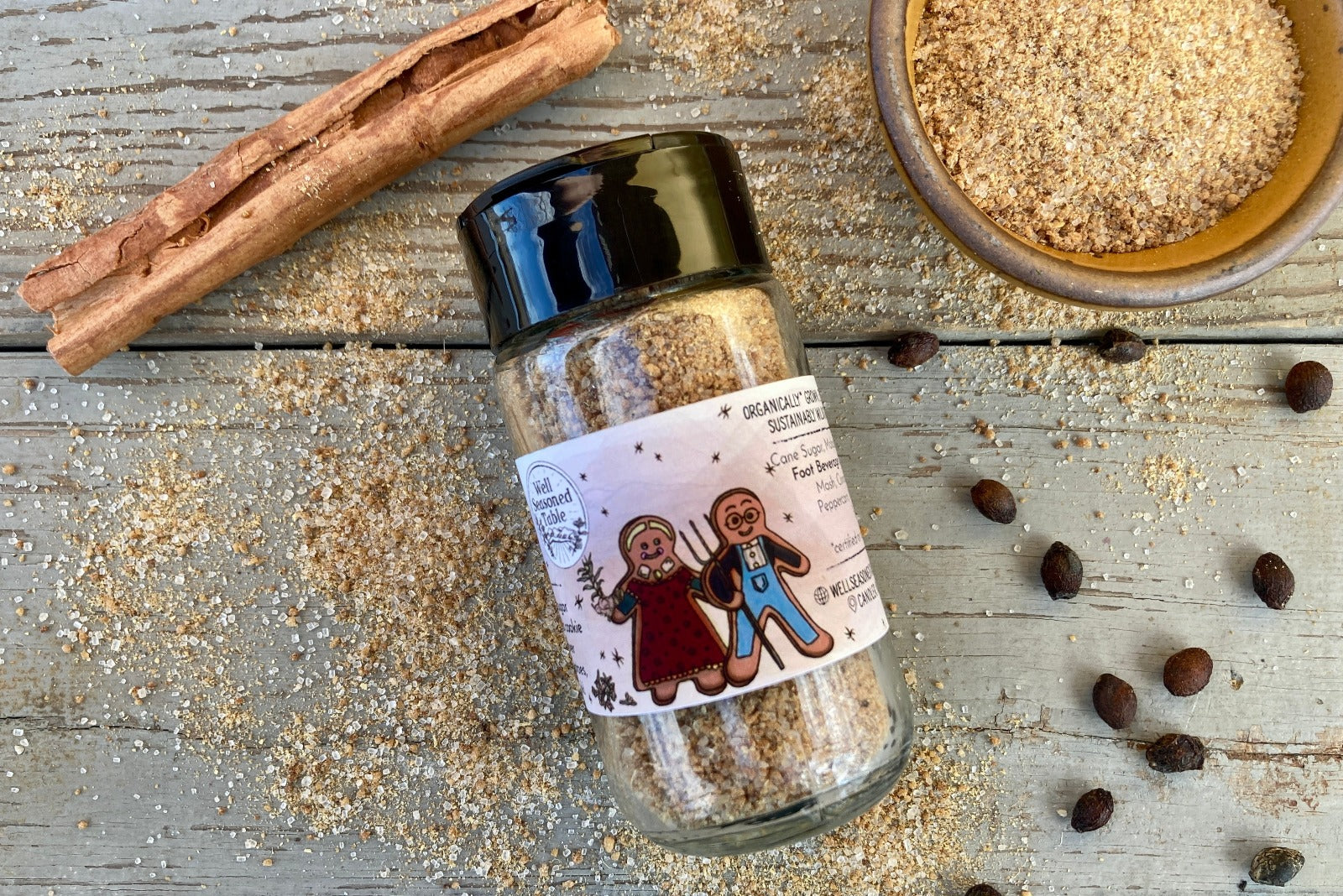 A glass shaker jar of Spiced Gingerbread Sugar Dust from Well Seasoned Table on a wooden background with a bowl of gingerbread sugar and a cinnamon stick.