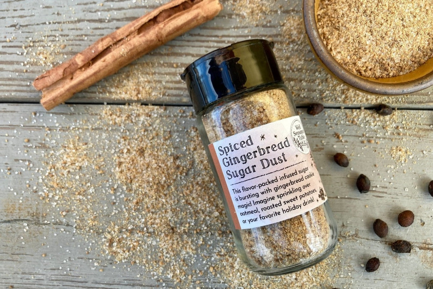 A glass shaker jar of Spiced Gingerbread Sugar Dust from Well Seasoned Table on a wooden background with a bowl of gingerbread sugar and a cinnamon stick.